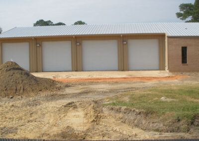 New garage structure with three doors and white metal siding on a dirt construction site.