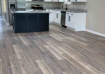 Open kitchen with white cabinets, dark blue island, granite countertops and wide-plank gray-brown vinyl flooring.